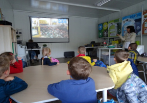 Dzieci siedzą przy stolikach i oglądają prezentację multimedialną.